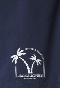 Marineblått stoff med et hvitt palmetrær grafikk med geometriske detaljer, og teksten "JACK & JONES ETABLERT 1990" nederst.