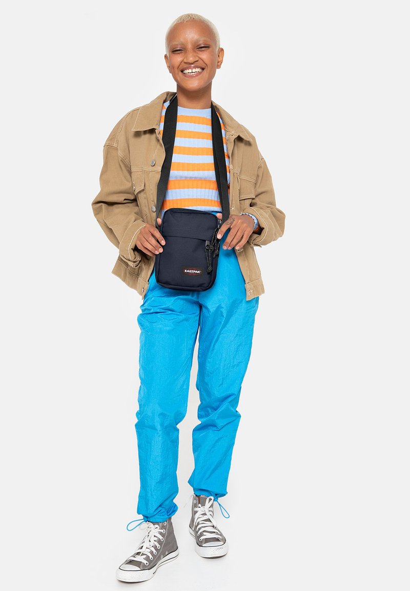 Borsa a tracolla nera con zip, giacca beige oversize, pantaloni blu e sneakers alte grigie. Camicia a righe arancione e blu.