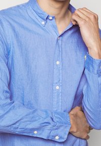 Light blue button-up shirt made of textured fabric, featuring a semi-collar, long sleeves, and white buttons. Subtle stitching detail visible.