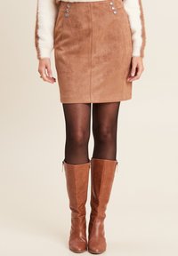 Frau trägt einen sandfarbenen Wildleder-Minirock, schwarze Strumpfhosen, braune kniehohe Lederstiefel und einen weißen, flauschigen Pullover mit braunen Streifen an den Ärmeln.