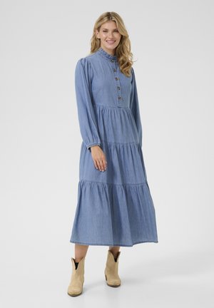 Blue denim maxi dress with a ruffled hem, button-up front, high neckline, and long sleeves. Worn with tan ankle boots.