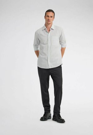 Uomo in piedi con le mani dietro la schiena, indossa una camicia a righe grigio chiaro con bottoni, pantaloni neri e stivali neri con lacci su sfondo semplice.