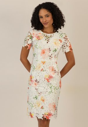 Frau mit lockigem Haar, die ein weißes knielanges Spitzenkleid mit floralem Muster und kurzen Ärmeln trägt, steht vor einem einfarbigen Hintergrund.