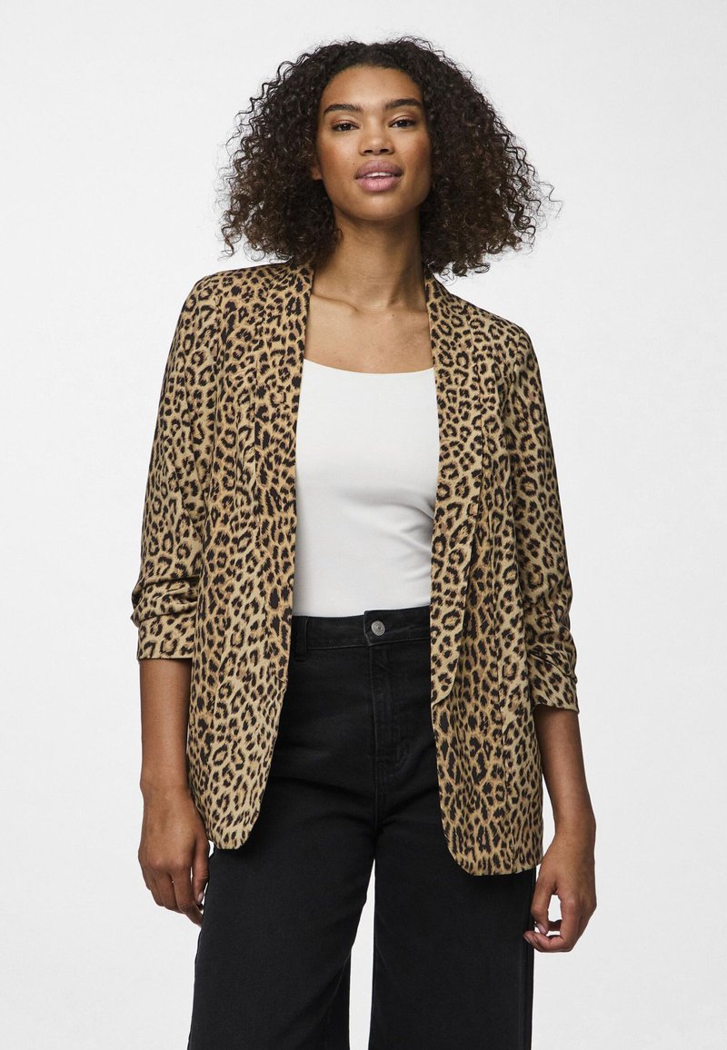 Femme avec des cheveux bouclés portant un blazer imprimé léopard sur un haut blanc et un pantalon noir taille haute, se tenant devant un fond uni.