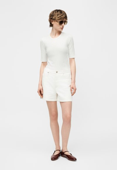 Γυναίκα που φοράει λευκό κοντομάνικο τοπ, λευκά σορτς, καφέ παπούτσια Mary Jane και γυαλιά ηλίου, στέκεται με τα χέρια χαλαρά δίπλα στο σώμα.