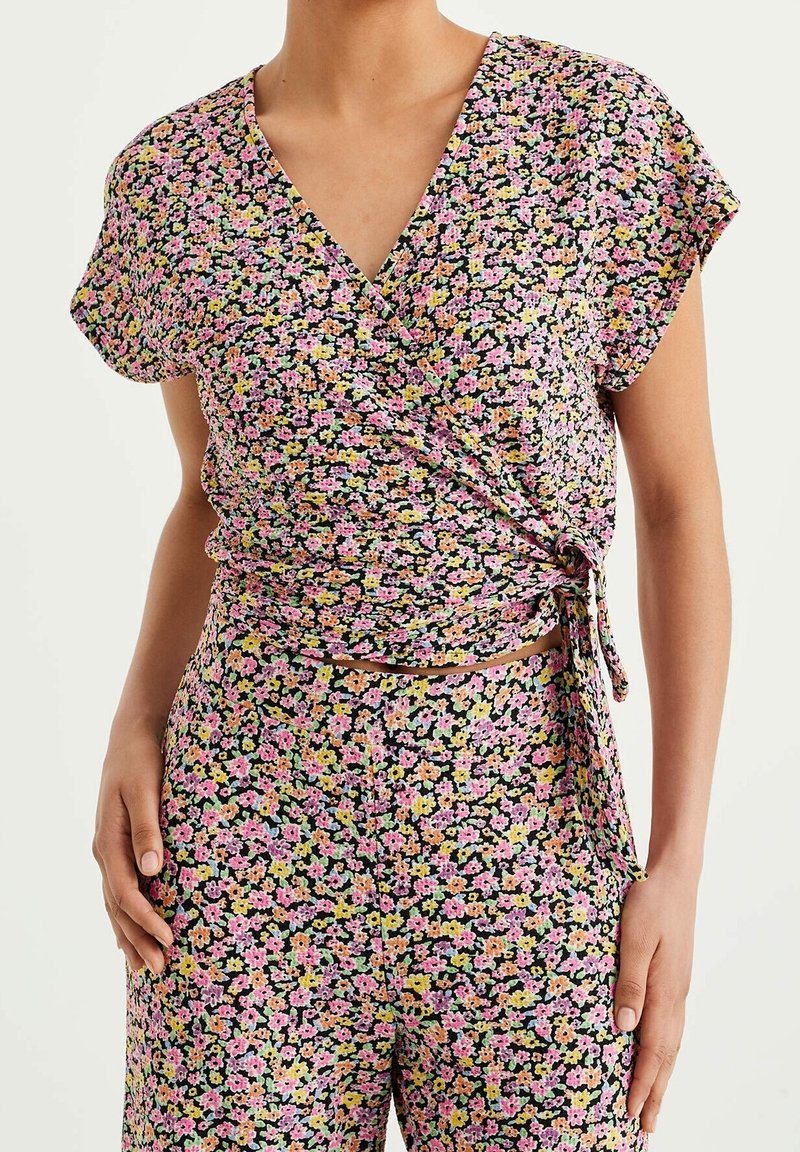 Femme portant un ensemble top cache-cœur à fleurs et pantalon assorti en rose, jaune et vert sur fond noir.