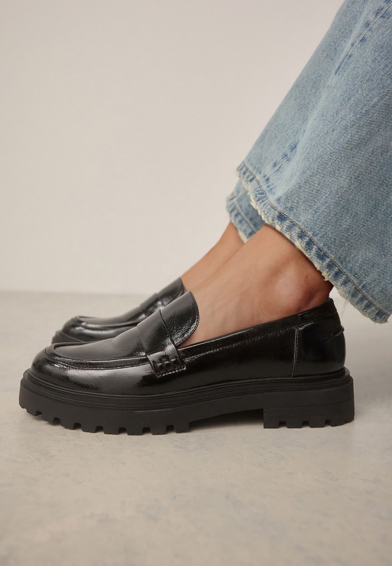 Mocassins en cuir noir brillant avec semelles épaisses à crampons portés avec un jean bleu clair à ourlet effiloché sur un fond de sol clair.