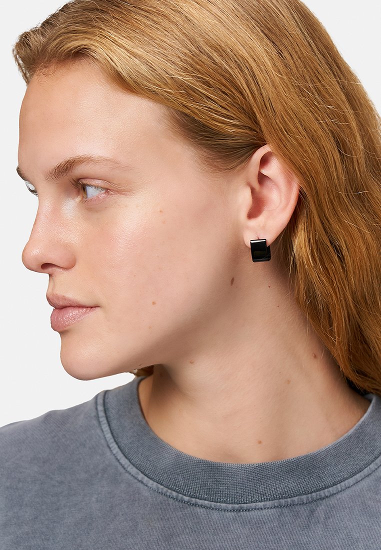 Schwarze quadratische Stud-Ohrringe mit metallischen Akzenten, im Ohr getragen. Das Model hat hellbraune Haare und trägt einen grauen Sweatshirt. Einfaches und geometrisches Design.