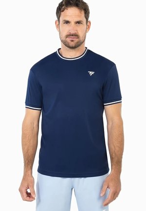 Hombre con una camisa de manga corta azul marino con revestimiento blanco en el cuello y las mangas, de pie frente a un fondo blanco.