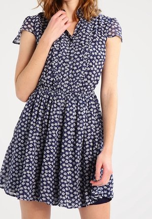 Vrouw draagt een marineblauwe jurk met korte mouwen, witte bloemenprint en gerimpelde kraag, staat met één hand bij haar nek.