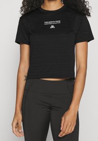 Svart croppad t-shirt i ett slätt tyg, med en vit logotyp "The North Face Mountain Athletics" på bröstet.