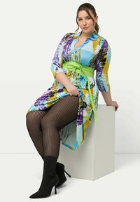 Abito stampato colorato con un motivo blu e giallo, cintura verde annodata, collant a strisce nere e stivaletti neri alla caviglia.