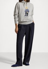 Sudadera de algodón gris con un gráfico de un oso que lleva un suéter azul, combinada con pantalones de pierna ancha azul marino y zapatillas azules.