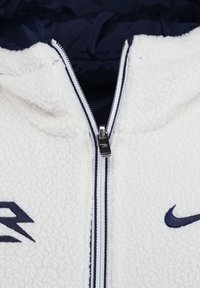 Veste en polaire blanche avec fermeture éclair, doublure marine et accents contrastants en marine. Présente une broderie de logo sur la poitrine et une fermeture éclair lisse.