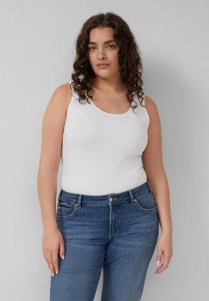 Vrouw met krullend bruin haar die een wit mouwloos topje en blauwe jeans draagt, staand tegen een grijze achtergrond.