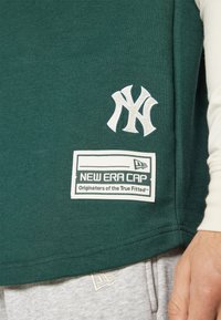 Tmavop green krátky rukáv tričko s bielym vyšívaným logom "NY" a bielou obdĺžnikovou nášivkou s nápisom "NEW ERA CAP".