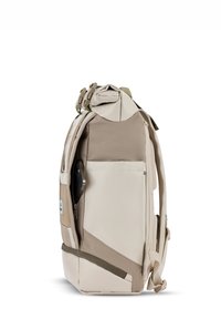 Beige színű hátizsák, hengeres tetővel, textúrált panelekkel, állítható pántokkal, elülső zsebbel, és egy látható okostelefonnal az oldalzsebében.