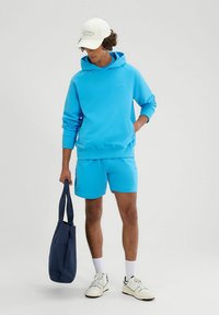 Completo di felpa blu e pantaloni corti realizzato in materiale morbido. Gli accessori includono un cappellino bianco e una borsa tote blu scuro. Le sneakers completano l'outfit.