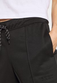 Černé jogger kalhoty z texturované látky, s páskem na šňůrku, bočními kapsami a vyraženým logem na levé nohavici.