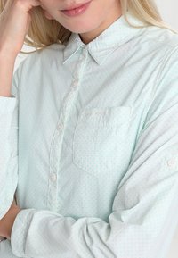 Blusa verde chiaro con un design a bottone, caratterizzata da piccoli motivi circolari, una tasca sul petto e maniche lunghe arrotolate.
