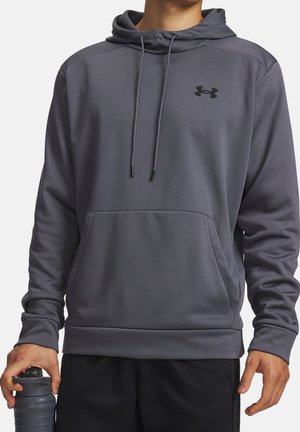 Homme portant un sweat à capuche gris Under Armour avec poche ventrale et cordons, tenant une bouteille d'eau grise dans la main gauche.