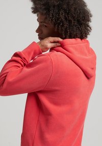 Felpa rossa con cappuccio a coulisse, polsini a costine e una texture morbida. Il retro è semplice, senza loghi o modelli visibili.