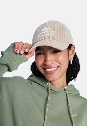 Beżowa czapka z sztruksu z białym haftowanym logo "ROXY"; model nosi zieloną bluzę z kapturem, lekko teksturowaną tkaninę, luźny krój.