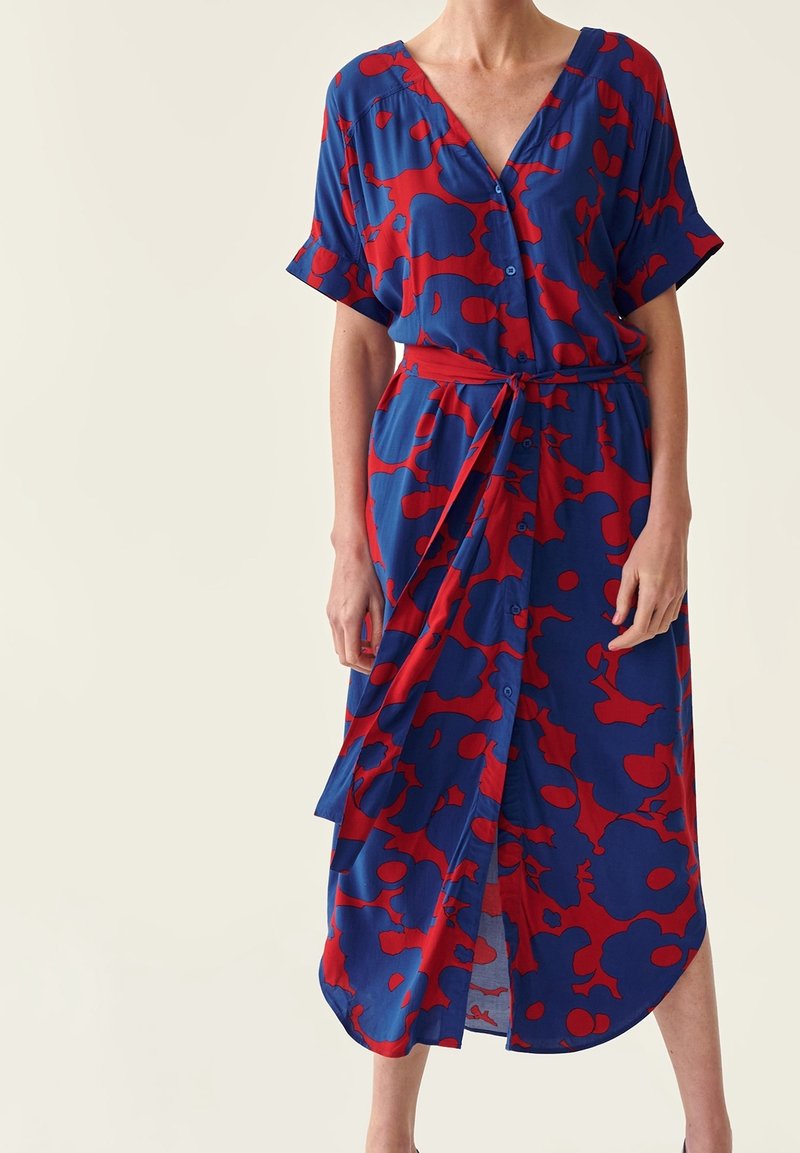 Femme portant une robe midi portefeuille à motifs bleus et rouges, manches courtes et décolleté en V, nouée à la taille avec une ceinture assortie.