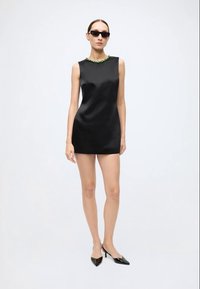 Black satin sleeveless dress with a round neckline adorned with green detailing, paired with black pointed heels and sunglasses.