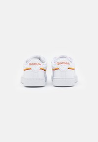 Ténis brancos da Reebok com pormenores em laranja e amarelo, material de couro liso, forma arredondada, solas planas e solas exteriores de borracha texturizada.