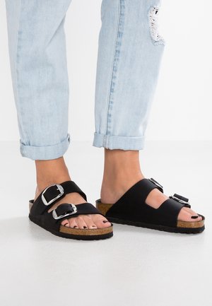 Black slide sandals with two adjustable metallic buckles. Cork footbed and rubber outsole. Worn with rolled-up light blue jeans.