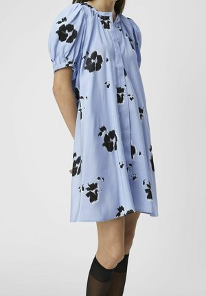 Robe bleue longueur genou avec imprimés floraux noirs, manches courtes bouffantes et bas noirs transparents montant jusqu'aux genoux sur une personne debout.