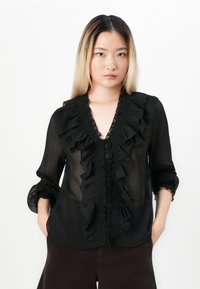 Femme aux longs cheveux ombrés portant une blouse noire transparente avec un devant et des manches volantés, se tenant avec les mains dans les poches devant un fond blanc.