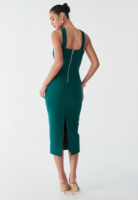 Vestido midi verde ajustado con tirantes gruesos, tela texturizada, cremallera trasera y abertura lateral en el dobladillo. Combinado con tacones de color claro.