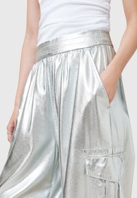 Personne portant un pantalon large argenté brillant avec une taille élastique, des poches latérales et une poche cargo, associé à un haut blanc sans manches.