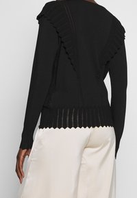 Pull en maille noire avec des accents froncés festonnés sur les épaules et une texture côtelée en bas, associé à un pantalon large crème.