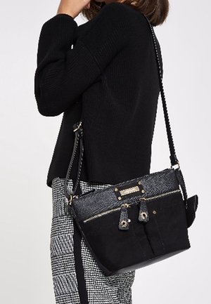 Kobieta w czarnym swetrze i kraciastych spodniach, nosząca czarną torbę na ramię z przednimi kieszeniami na zamek i teksturowanym górnym panelem.