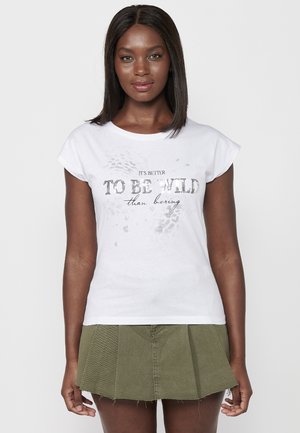 T-shirt blanc à manches courtes avec une impression graphique disant "Il vaut mieux être sauvage que ennuyeux", associé à une jupe en denim plissée verte.