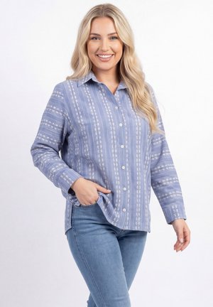Smiling woman with long blonde hair wearing a blue patterned button-up shirt and light blue jeans, hand in pocket, against white background.