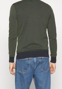 Neulottu vihreä neulepaita, jossa on teksturoitu kuvio ja resorit tummansinisinä hihansuissa, esitetty takaa sinisten farkkujen päällä.