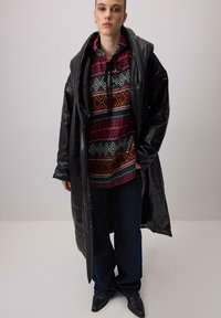 Sort læder lang frakke over en farverig mønstret hættejakke med geometriske designs i rød, grøn og hvid, parret med mørke vide jeans.