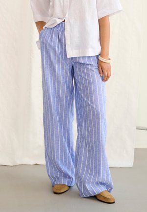 Osoba nosiaca voľné, svetlomodré nohavice s bielymi vertikálnymi pruhmi, bielu košeľu, béžové topánky a svetlé náramky, stojaca s jednou rukou v vrecku.