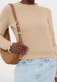 Femme portant un pull beige et un jean bleu clair, tenant un sac à bandoulière en daim brun avec des détails de boucle dorée.