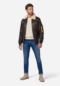 Braune Lederbomberjacke mit Lammfellkragen, Reißverschlusstaschen, gerippten Bündchen, kombiniert mit hellblauen Jeans und beigen Stiefeletten.