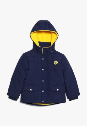 Veste matelassée bleu marine avec doublure polaire jaune, capuche amovible, boutons-pression à l’avant, deux poches à rabat et un patch rond jaune sur la poitrine.