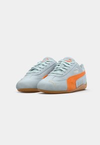 Tênis de camurça azul claro com um detalhe laranja, apresentando uma sola de borracha natural, atacadores e um logotipo branco na lateral.
