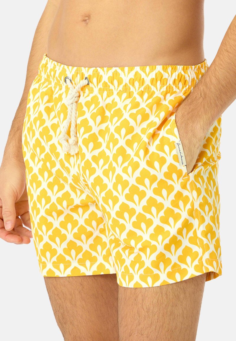 Homme portant un short de bain à motifs jaunes et blancs avec une taille coulissante, main dans la poche droite, sur un fond uni.