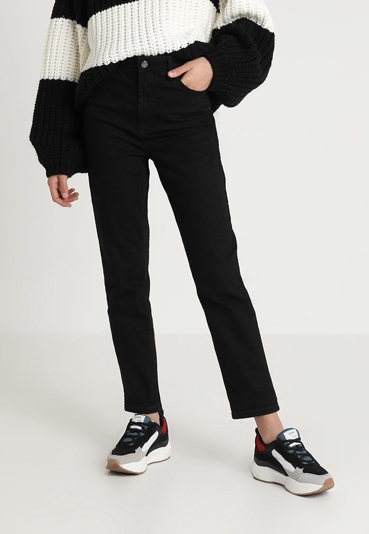 Personne portant un jean noir, un pull en tricot rayé noir et blanc, et des baskets noires, blanches et rouges, debout avec une main dans la poche.