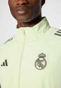 Zelena jakna na zip s teksturiranom površinom, crnim Adidas trakama i izvezenim logom Real Madrida na prsima.