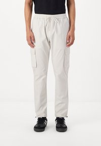 Hellbeige Cargo-Hosen mit elastischem Bund, Kordelzugverschluss, zwei seitlichen Taschen und glatter Textur, kombiniert mit schwarzen Sneakers.